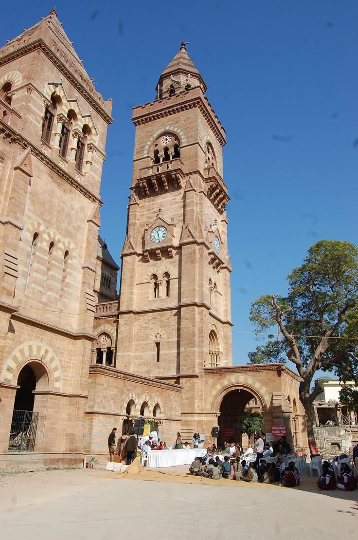 two tall towers with clocks on them in front of a building and people sitting at tables