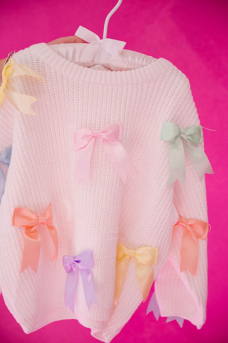 a white sweater with colorful bows hanging from it's hanger against a pink background