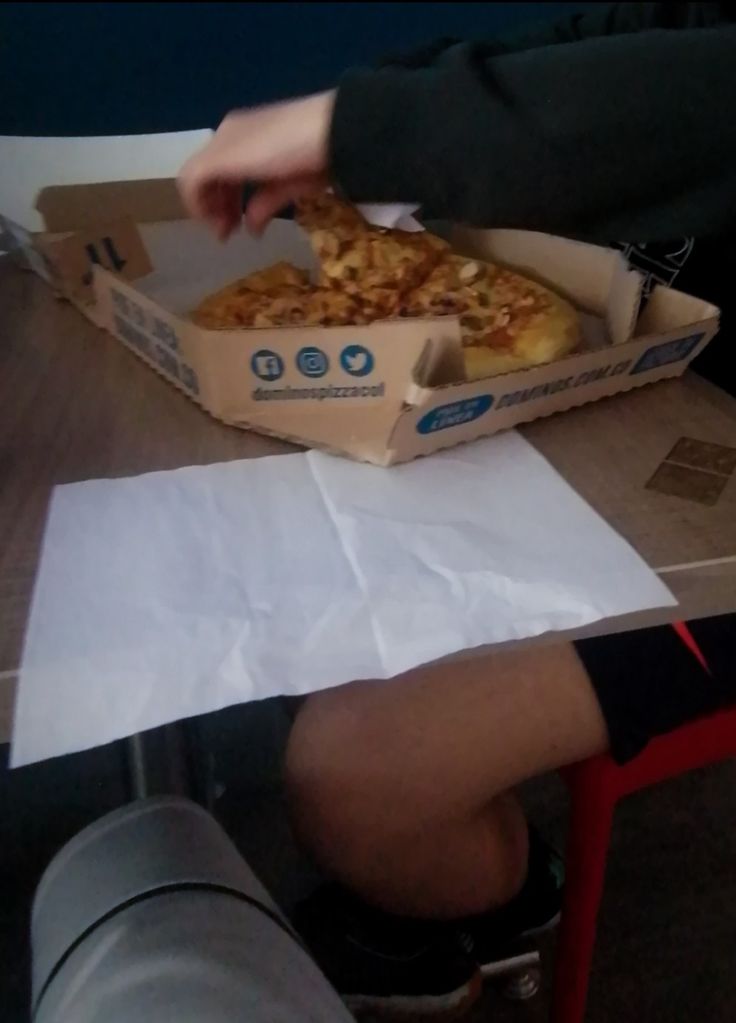 a person sitting at a table with a box of pizza in front of him and one hand reaching for it