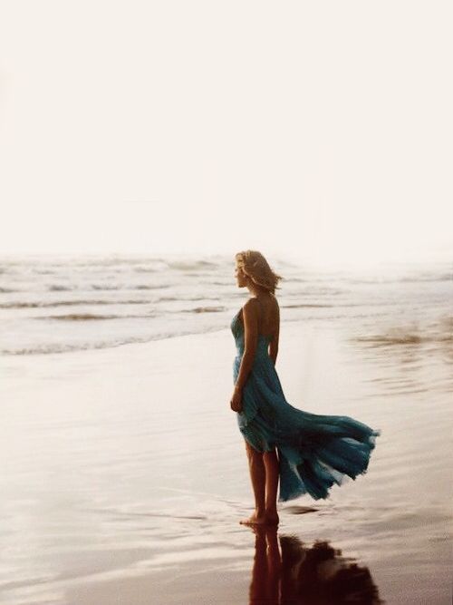 a woman in a blue dress standing on the beach looking out at the water and waves