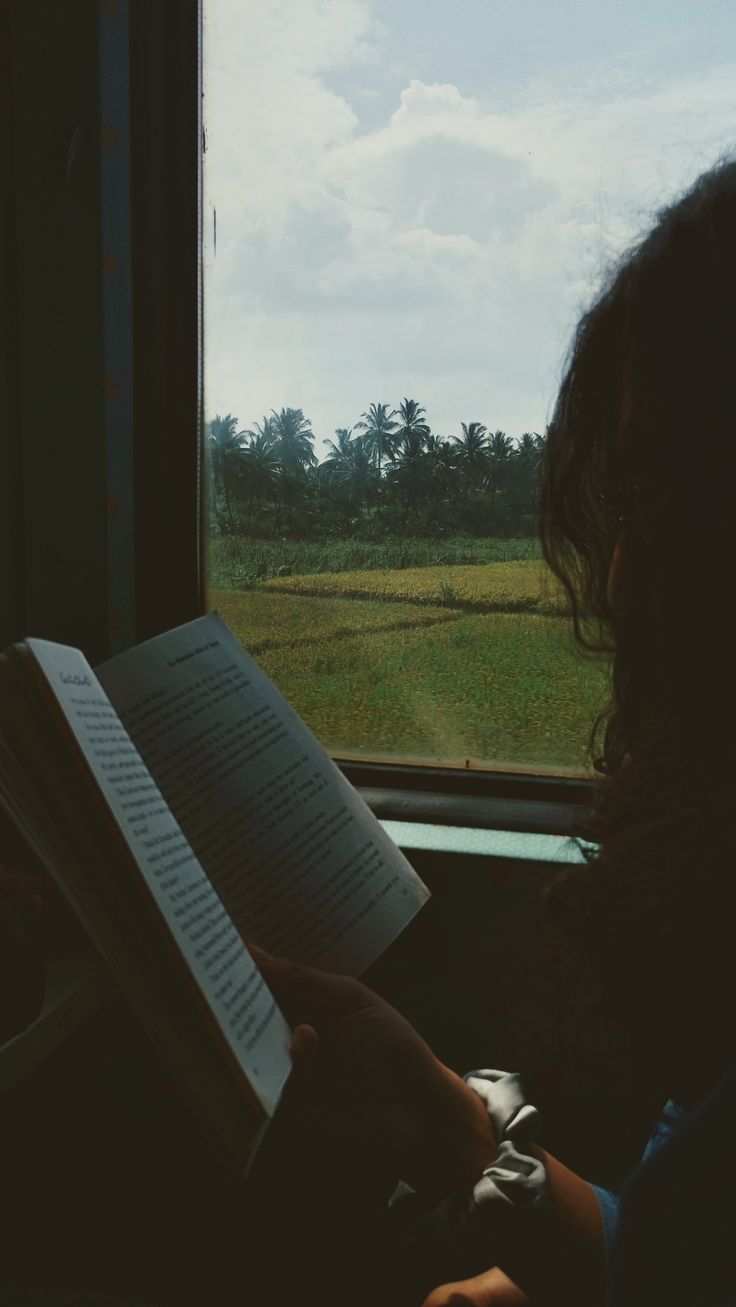 a person sitting on a train reading a book next to a window with grass and trees in the background