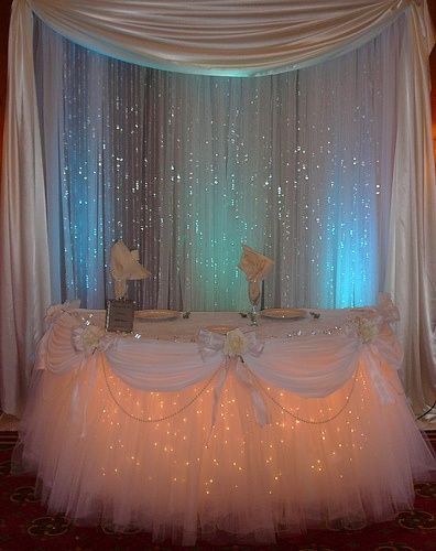 a table covered in white tulle with lights on it and flowers at the top