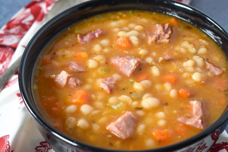 a bowl of soup with ham, beans and carrots on a red and white napkin