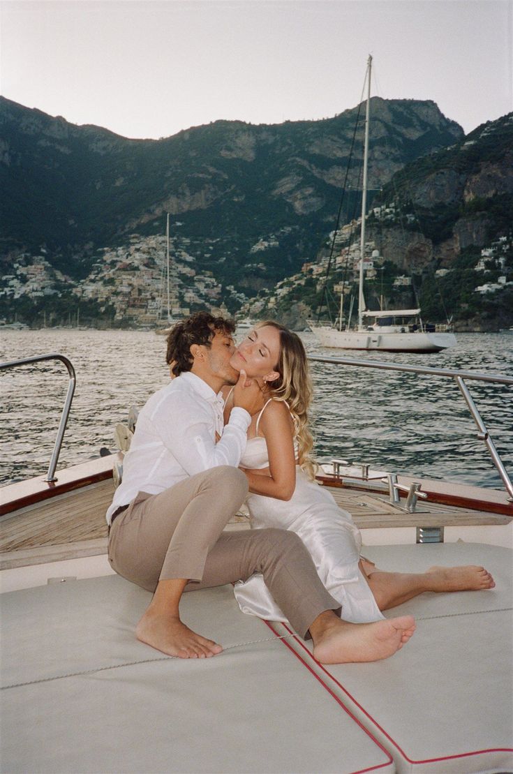 a man and woman sitting on the back of a boat kissing in front of mountains