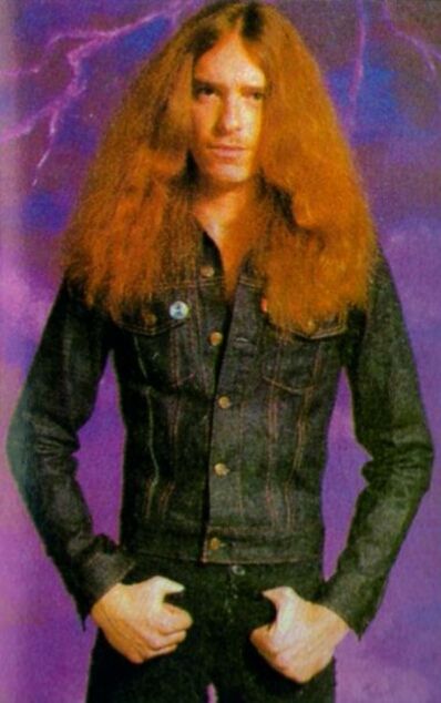 an image of a man with long red hair wearing black jeans and denim jacket standing in front of a purple background