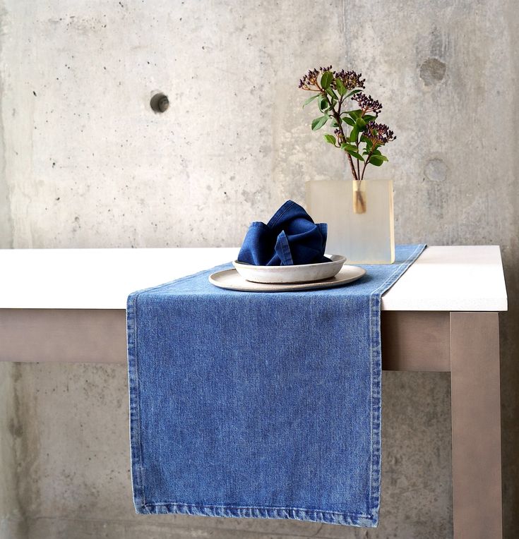 a blue table runner with a white plate and flower in a vase on the edge