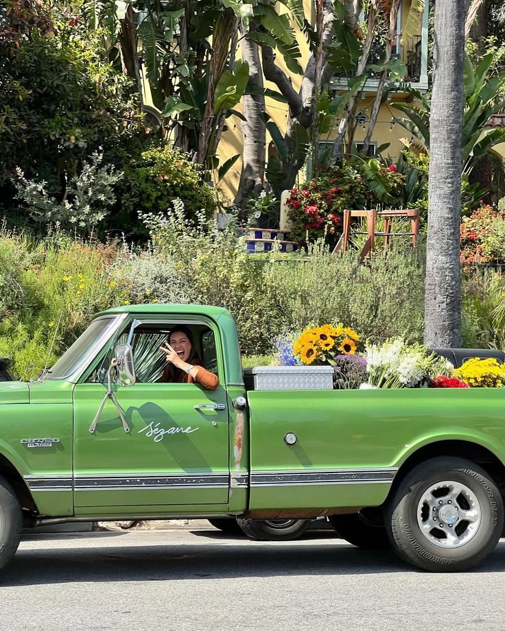 a green pick up truck with flowers in the bed driving down a street next to palm trees