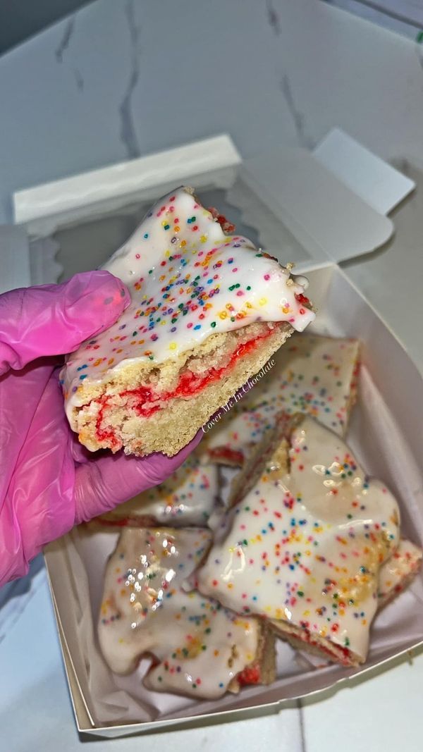 a person wearing pink gloves holds up a box of cookies with white frosting and sprinkles