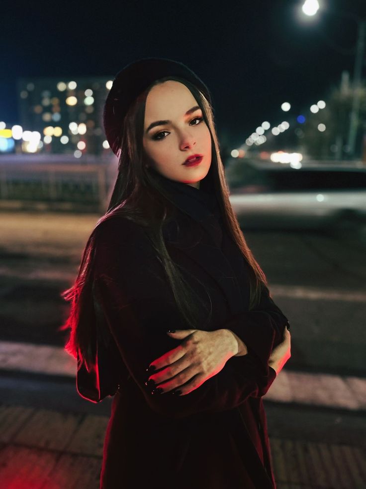 a woman standing on the street at night with her arms crossed and looking off to the side