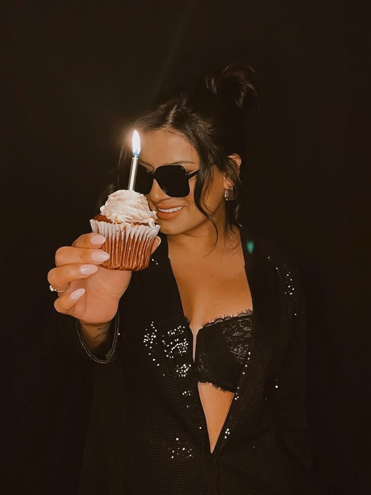 a woman wearing sunglasses holding a cupcake with a candle in it's mouth