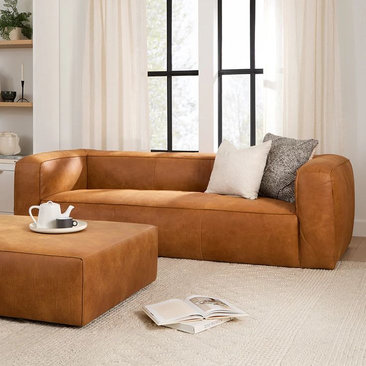 a brown leather couch sitting in front of a window next to a coffee table and ottoman