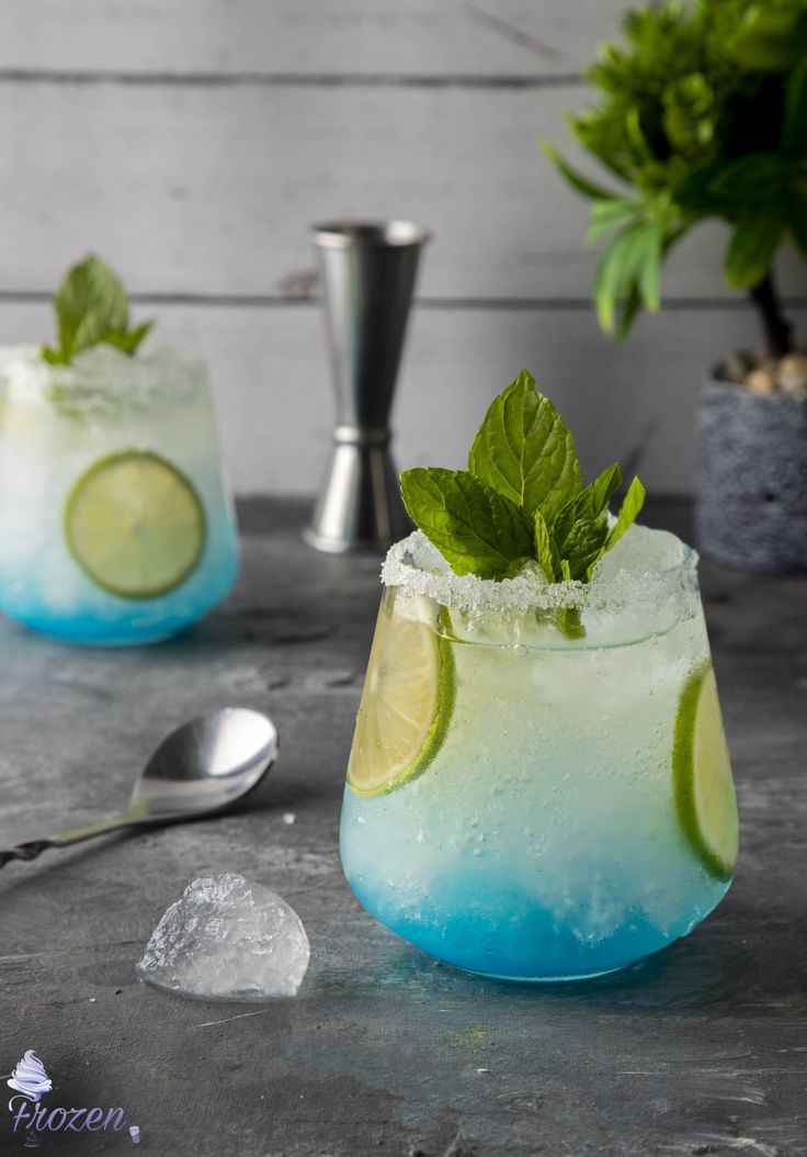 two glasses filled with blue and green drinks next to a silver spoon on top of a table