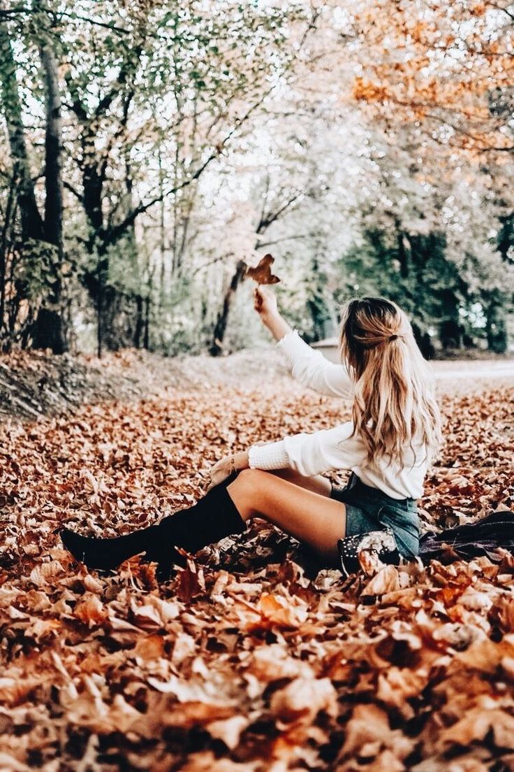 a woman sitting on the ground surrounded by fallen leaves and trees with her arms in the air