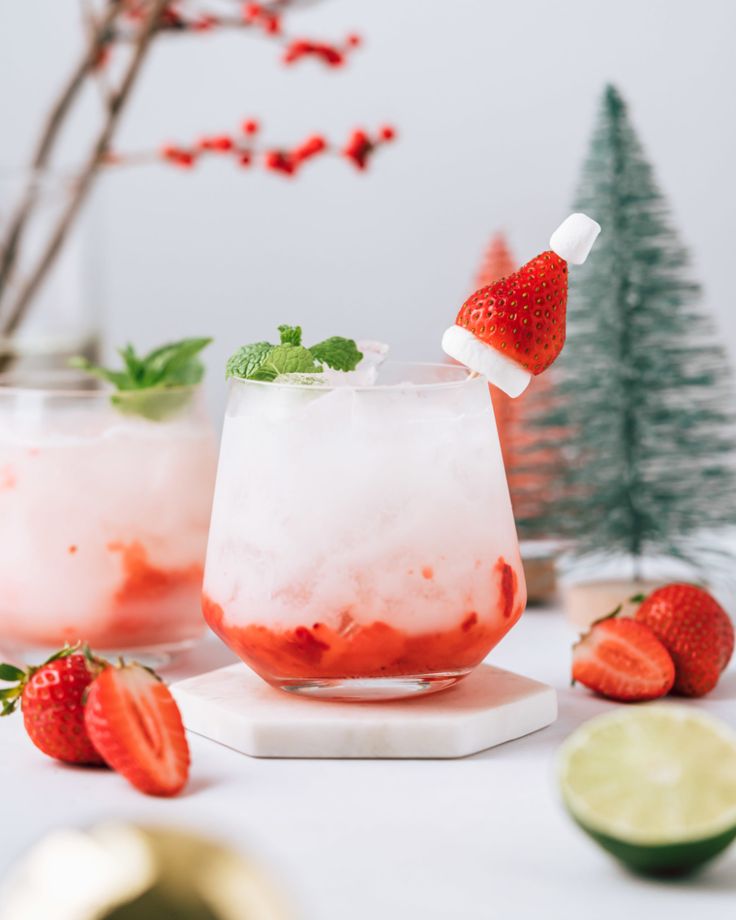 two glasses filled with ice and strawberries next to limes on a white table