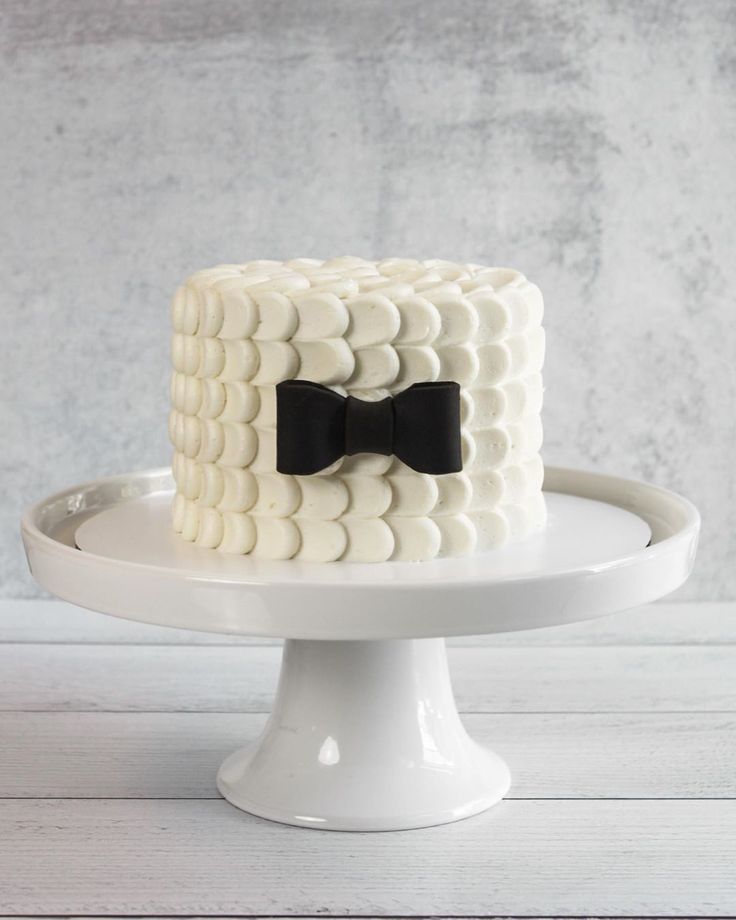 a white cake with a black bow tie on it sitting on a plate in front of a gray wall