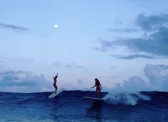 two people on surfboards riding the waves in the ocean at dusk with full moon behind them