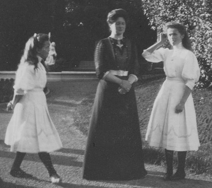 three women in long dresses standing next to each other on a sidewalk with trees and bushes behind them