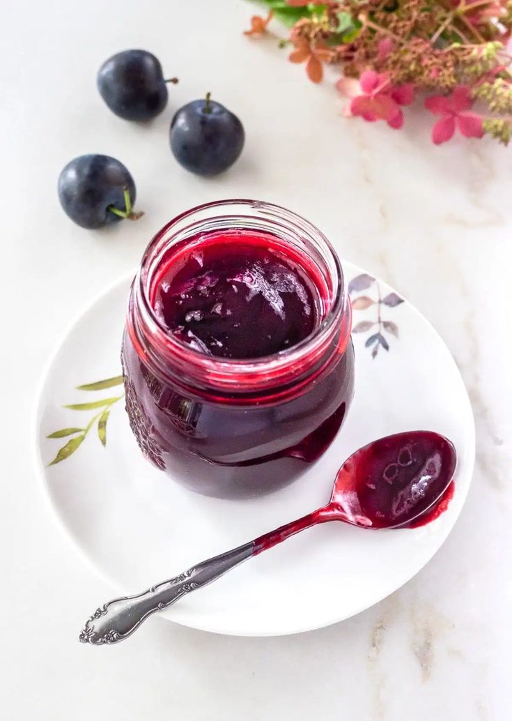 a jar of blueberry jam on a white plate with spoons next to it