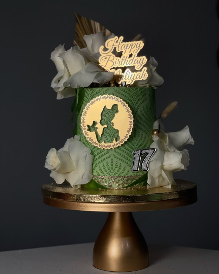 a green birthday cake with white flowers on a gold plated stand and a happy birthday sign