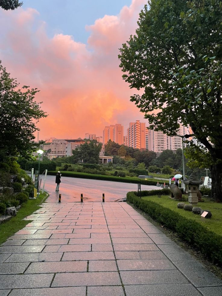 a person walking down a sidewalk in front of some trees and buildings with pink clouds