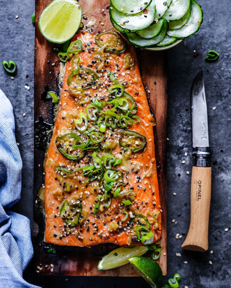 a piece of salmon on a cutting board with sliced cucumbers and limes