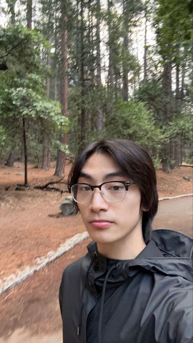 a young man wearing glasses taking a selfie in the woods with his cell phone