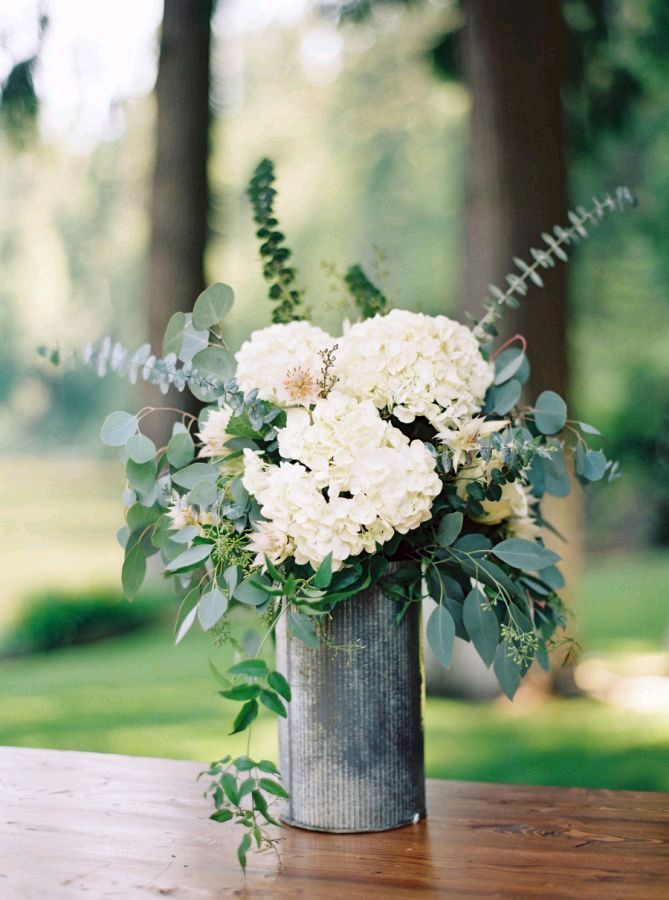 a vase filled with white flowers sitting on top of a wooden table covered in greenery