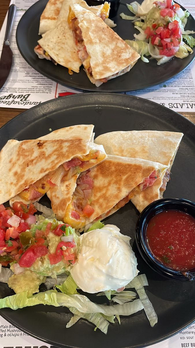 two black plates topped with quesadillas and salad next to a bowl of salsa