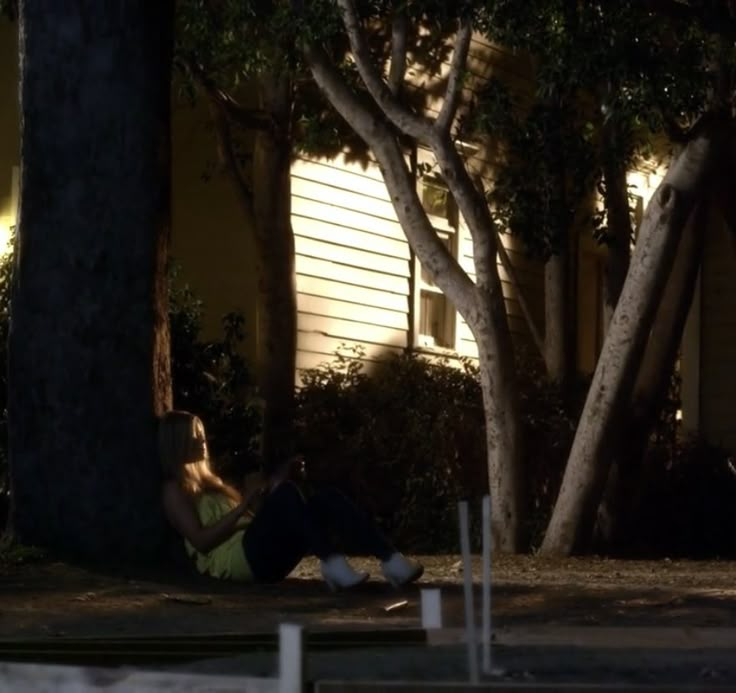 a woman sitting on the ground in front of a house at night with her cell phone up to her ear