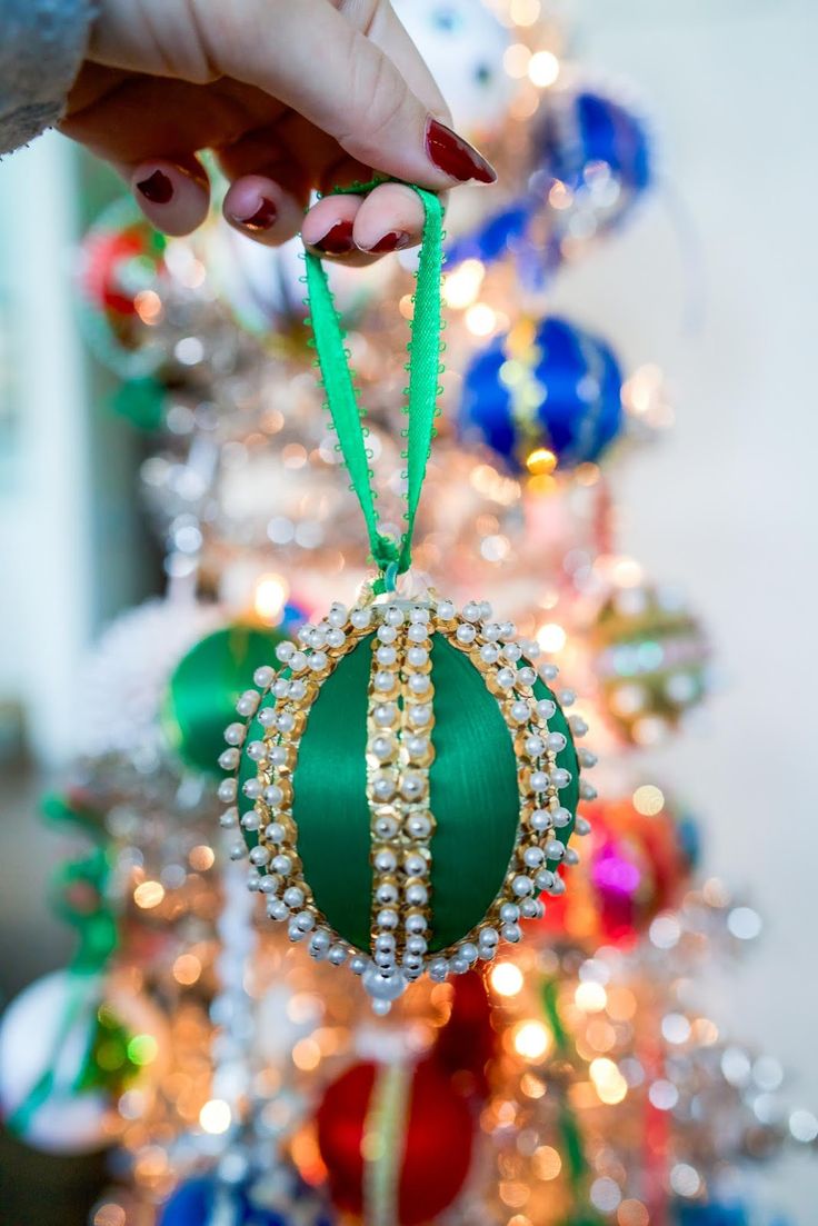 a hand holding a christmas ornament in front of a tree with ornaments on it