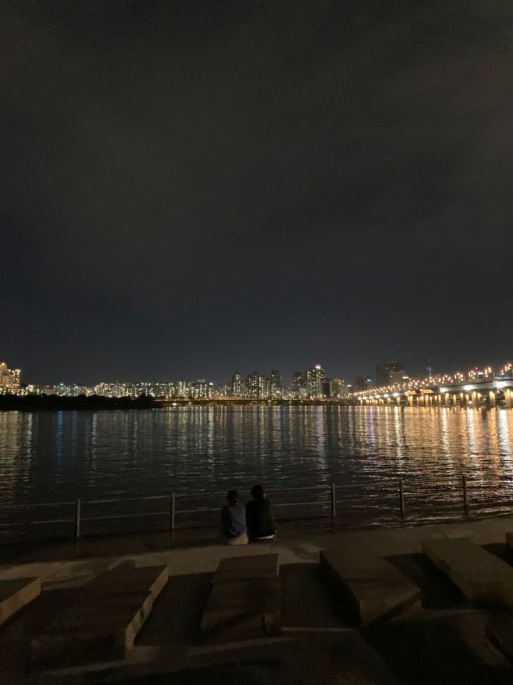 two people sitting on the edge of a body of water at night with city lights in the background