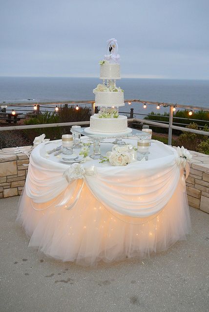 a wedding cake on top of a table with white flowers and lights in front of the ocean