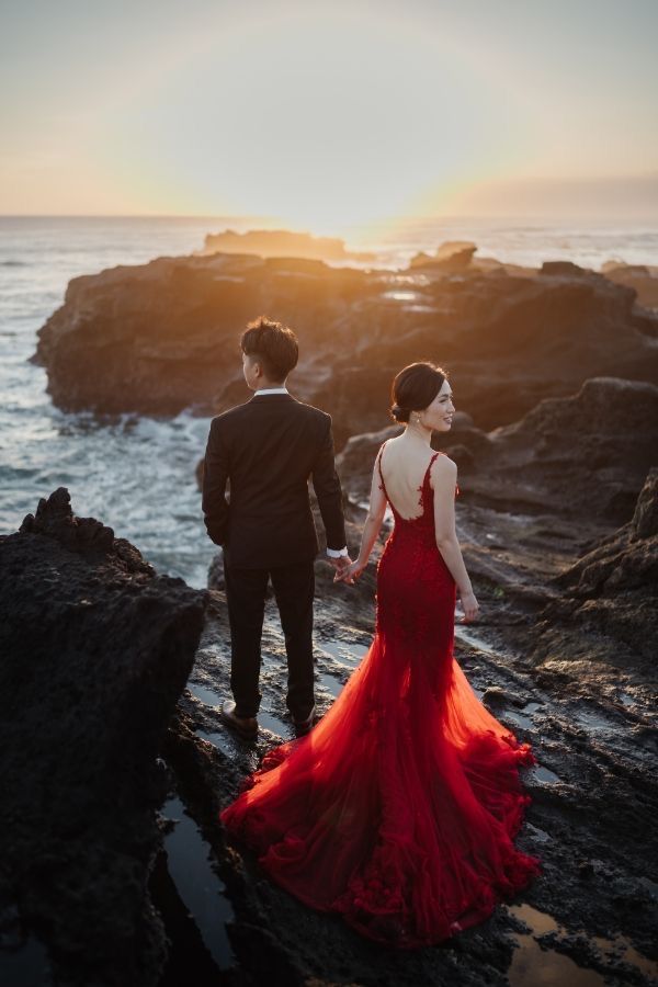 a man and woman in formal wear standing on rocks near the ocean at sunset, holding hands