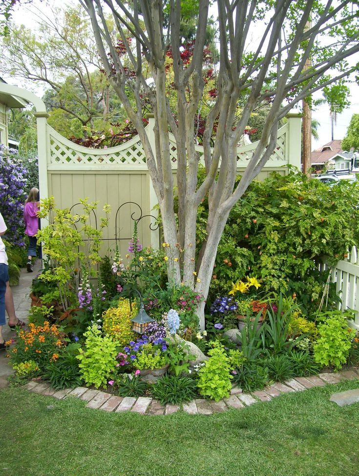 a woman standing in front of a tree surrounded by flowers and plants on the grass