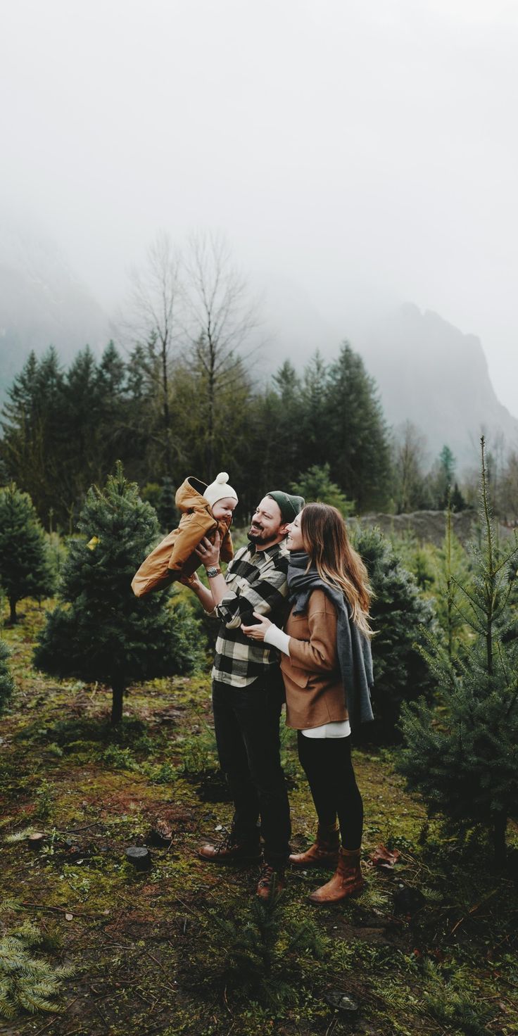 a man and woman standing in the middle of a christmas tree farm holding an infant