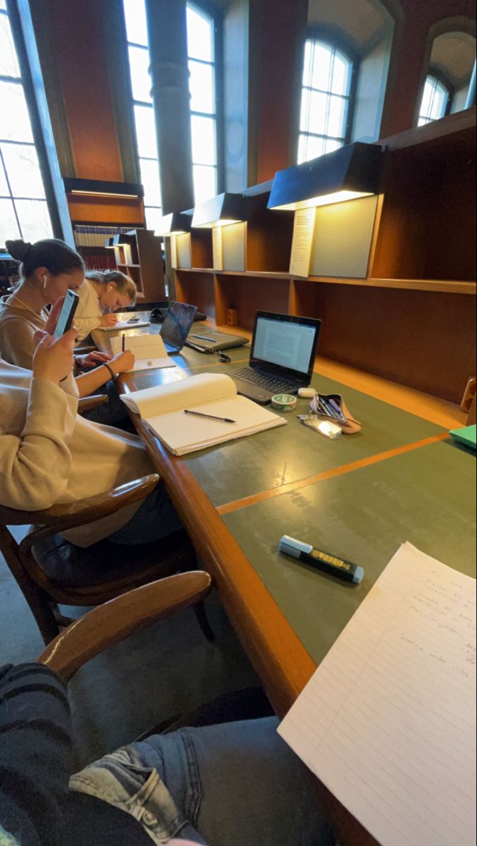 two people sitting at a long table with laptops and papers on it, while one person holds a cell phone up to their ear