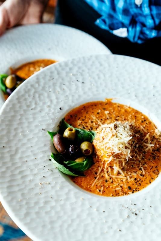 a white plate topped with a bowl of soup covered in cheese and olives on top of a blue table cloth