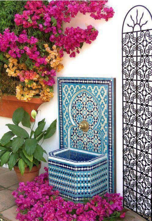 colorful flowers are blooming in front of a white wall with a fountain and potted plants