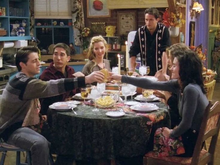 friends sitting around a table with food and drinks in the kitchen, one person reaching out to shake hands