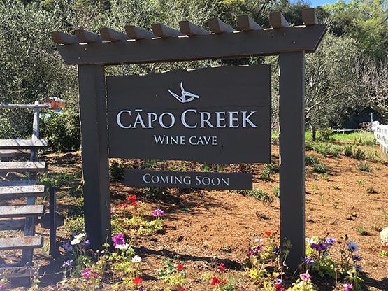 a sign for capo creek winery cave in the middle of a field with flowers around it