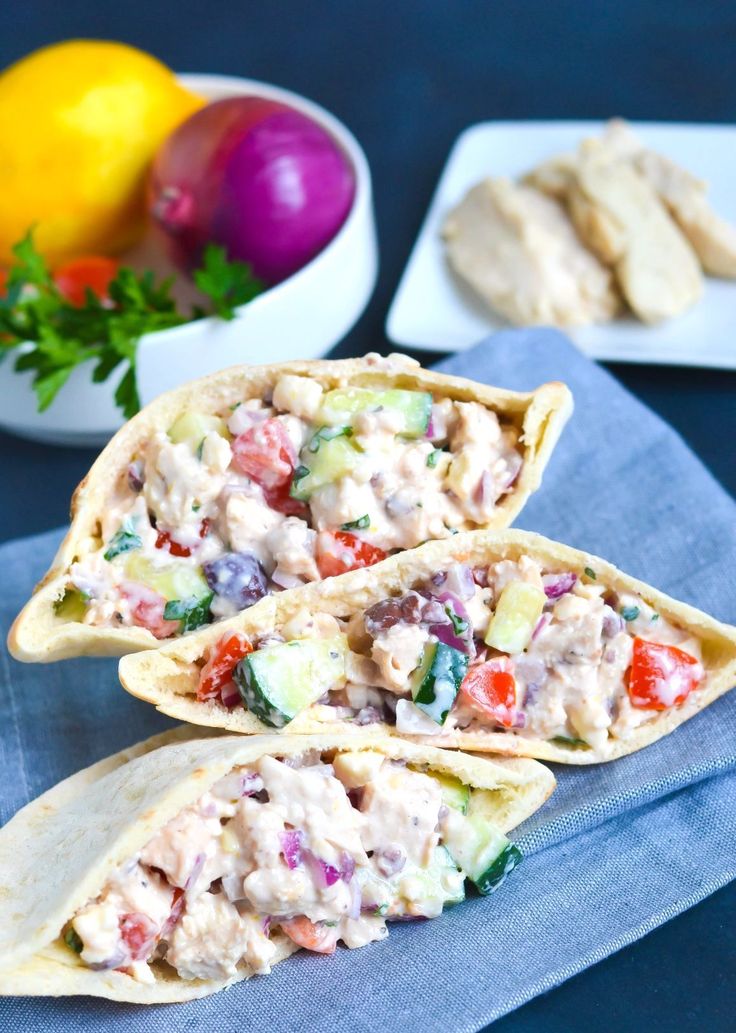 two pita breads filled with chicken salad on top of a blue napkin next to bowls of vegetables