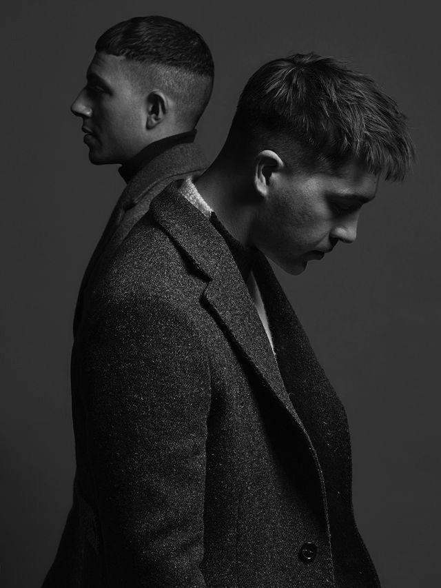 two men standing next to each other in front of a dark background, one wearing a coat