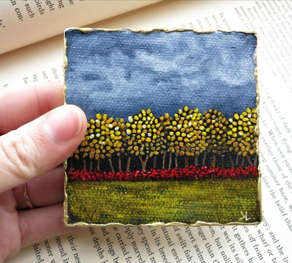 a hand holding up a small painting on top of an open book with trees in the background