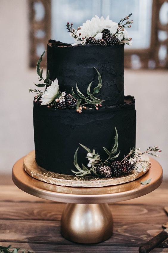 a three tiered black cake with white flowers and greenery on top sits on a gold plate