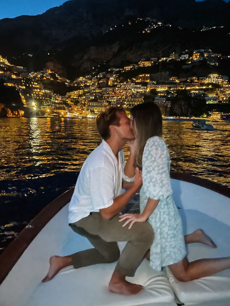 a man and woman kissing on the back of a boat in front of a city at night