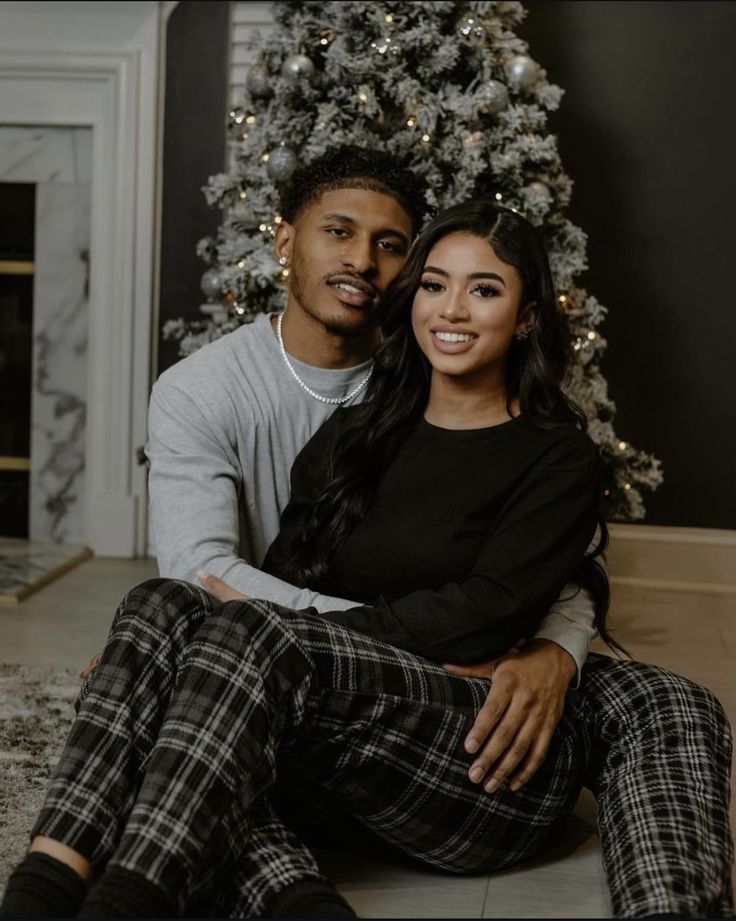 a man and woman sitting in front of a christmas tree with their arms around each other