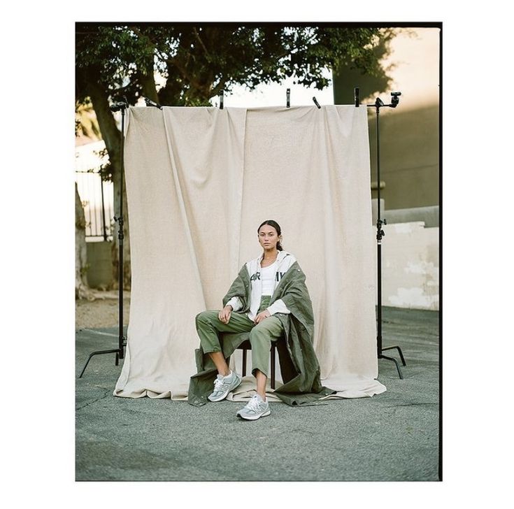 a woman is sitting on a chair in front of a white backdrop and some trees