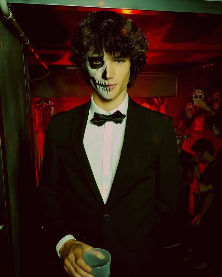 a man in a tuxedo and skull makeup is holding a cup while wearing a black suit