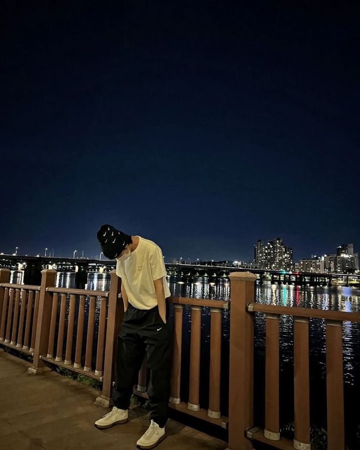 a man standing on top of a wooden fence next to the water at night with city lights in the background