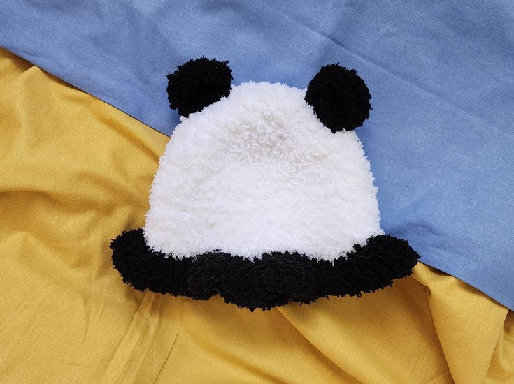 a panda bear hat laying on top of a blue and yellow bed spread with it's eyes closed